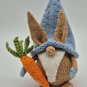 Könnte beinhalten: Ein gestrickter Gnom mit braunen Hasenohren, einer blauen Mütze und einem weißen Bart, der eine Karotte hält.