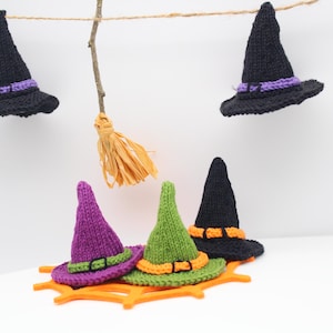 May include: A collection of handmade Halloween decorations featuring miniature witch hats in black, purple, and green, with orange and purple bands. A small broom hangs from a twig. The hats are displayed on a white surface.