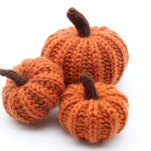 May include: Three knitted pumpkins in shades of orange, each with a brown stem. The pumpkins are of different sizes and are displayed against a white backdrop. The knitted texture is clearly visible, evoking a seasonal autumn theme.
