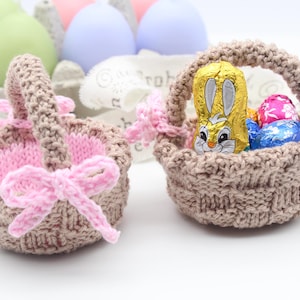 May include: Two small, hand-knitted Easter baskets in a light brown colour with pink accents and bows. One basket contains a gold foil-wrapped chocolate bunny and colourful Easter eggs. A carton of pastel-coloured eggs is in the background.