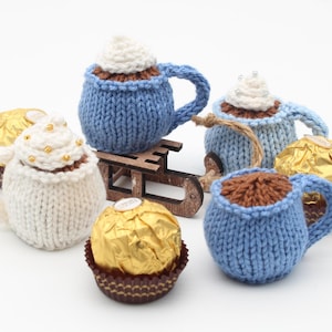 Könnte beinhalten: Fünf gestrickte Tassen in Form von heißer Schokolade und Schokoladenbonbons. Die Tassen sind blau und weiß mit einer gestrickten Textur. Die Bonbons sind in goldene Folie verpackt.