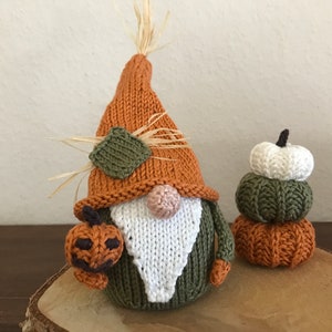 May include: A knitted gnome with a brown hat, green beard, and white face. The gnome is holding a small orange pumpkin. There is a stack of three knitted pumpkins behind the gnome, with the top pumpkin being white, the middle pumpkin being green, and the bottom pumpkin being orange.
