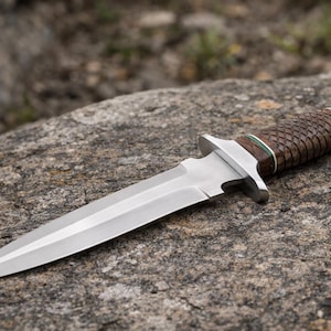 May include: A stainless steel dagger with a double-edged blade. The handle is wrapped in a brown, woven material with a silver bolster and a green accent. The knife is resting on a gray rock.