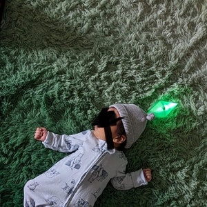 May include: A baby wearing a grey knit hat with a green light-up toy attached to it. The baby is lying on a green shag rug.