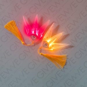May include: Two illuminated, crown-shaped hair accessories with attached yellow tassels. One glows pink, the other orange, each with an on/off button. The background is a light gray with the word "GIVE" repeated.