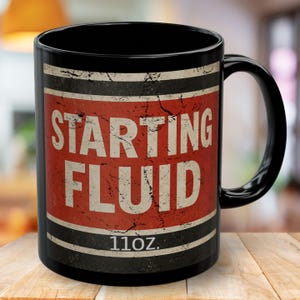 以下が含まれることがあります： 「STARTING FLUID」と書かれた、赤と白のラベルが付いた黒いセラミックマグ。マグカップの容量は312mlで、黒いハンドルが付いています。ヴィンテージ感のあるインダストリアルなデザインです。
