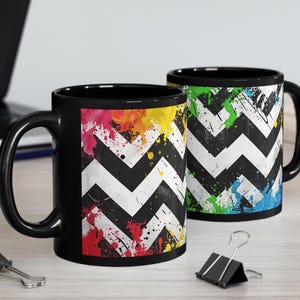 May include: Two black ceramic mugs with a white and black chevron pattern. One mug features colorful paint splatters, while the other has green, blue, and yellow splatters. Both mugs have black handles.