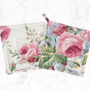 May include: Two square pot holders with floral rose patterns. One features a white background with pink roses and green leaves, the other has a similar design on a light blue background. Both have a loop for hanging.
