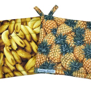 May include: Two square pot holders. One features a pattern of bananas, the other pineapples. Both have a quilted design and a loop for hanging. The pineapple pot holder has a small label that reads "Audrey Belisle".