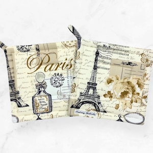 May include: Two square fabric pot holders featuring a cream background with a vintage Paris theme. The design includes black Eiffel Tower illustrations, a perfume bottle, script text, postmarks, and floral accents. Each holder has a small corner loop.
