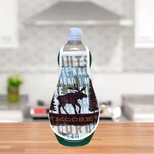 May include: A clear liquid soap dispenser with a decorative cover featuring a moose silhouette, trees, and the words "Nuts are natural, hear 1 more, moose lures". The cover has a green trim.