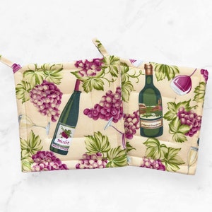 May include: Two beige quilted pot holders with a wine theme. Each pot holder features images of wine bottles, purple grapes, and green leaves. One bottle reads "Merlot", the other "Cabernet Sauvignon".
