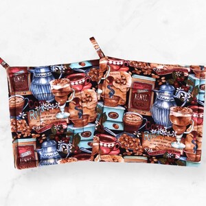 May include: Two square pot holders with a coffee-themed pattern. The fabric features coffee cups, coffee beans, and coffee pots in shades of brown, blue, and red on a black background. Each pot holder has a loop for hanging.