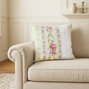 May include: A square throw pillow featuring a vertical striped pattern with alternating bands of pale yellow, light blue floral vines, and a central column of pink roses and green leaves on a cream-colored background with a white side panel.