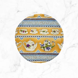 May include: Round, patterned fabric featuring a repeating design of teacups and teapots. The background is yellow with blue and white accents. The teacups and teapots are white with purple and green floral details. A decorative item for the kitchen.