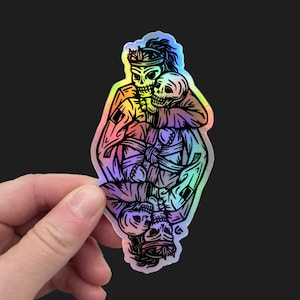 May include: Holographic sticker featuring a black and rainbow illustration of two skeletons embracing. One skeleton wears a crown. The sticker has a glossy finish and is held against a black background.
