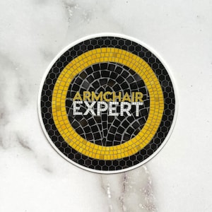 May include: A round sticker with a black and gold mosaic design. The text "ARMCHAIR EXPERT" is printed in white on the sticker.