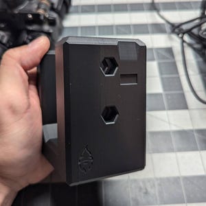 May include: A black, rectangular device featuring two hexagonal holes and a rectangular slot. The device has a small emblem at the bottom. A night vision device is visible in the background. The device is held in a hand.