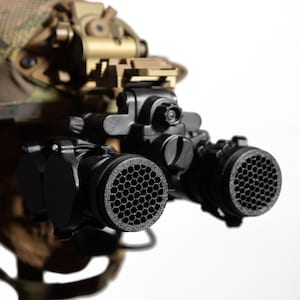 Puede incluir: Primer plano de gafas de visión nocturna negras con un patrón de panal en las lentes. Las gafas están montadas en un casco de color canela y camuflaje. La imagen está sobre un fondo blanco.