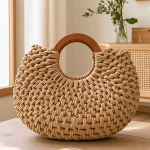 May include: A crescent-shaped handbag crafted from thick, beige fabric cord in a dense, woven pattern. The bag features a circular, polished wooden handle integrated into the top center, creating a distinct, rounded silhouette with a textured finish.