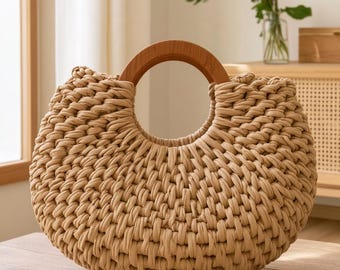 May include: A crescent-shaped handbag crafted from thick, beige fabric cord in a dense, woven pattern. The bag features a circular, polished wooden handle integrated into the top center, creating a distinct, rounded silhouette with a textured finish.