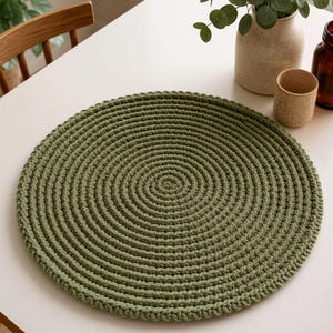 May include: A round, olive green crocheted placemat featuring a dense, concentric spiral stitch pattern. The placemat is crafted from thick yarn, creating a textured, circular surface with a consistent, uniform weave throughout the entire design.