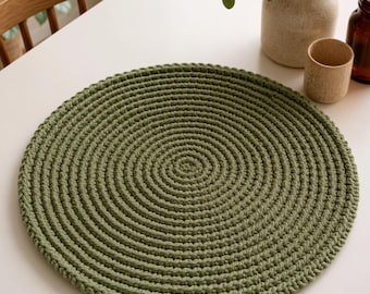 Puede incluir: Un mantel individual redondo de ganchillo en color verde oliva con un patrón de puntada en espiral concéntrica densa. El mantel está hecho de hilo grueso, creando una superficie circular texturizada con un tejido uniforme y consistente.