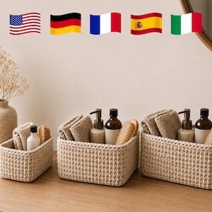 May include: Three rectangular beige woven storage baskets containing folded tan towels, brown pump bottles, and wooden brushes. The baskets feature a thick, textured knit pattern and are arranged in a row on a wooden surface with a vase nearby.