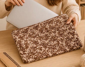 Puede incluir: Una funda rectangular para portátil con un patrón de ganchillo texturizado de dos tonos en tonos alternos de marrón oscuro y crema. La funda está construida con un diseño de punto denso y se muestra conteniendo un ordenador portátil plateado.