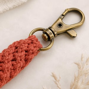 May include: A terracotta-colored macrame lanyard featuring a braided cord design attached to a swivel lobster clasp made of antique brass-toned metal. The cord is constructed from thick, textured cotton-like yarn in a deep, earthy orange hue.