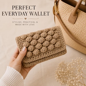 Pode incluir: Uma carteira de crochê bege com um padrão de pontos texturizados na aba frontal. O texto na imagem diz: PERFECT EVERYDAY WALLET, STYLISH, PRACTICAL & MADE WITH LOVE. Uma bolsa de palha trançada e um pequeno ramo de flores brancas são visíveis.