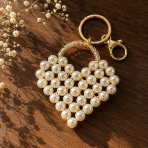 May include: A heart-shaped keychain constructed from cream-colored faux pearls arranged in a grid pattern. The top of the heart features a clear beaded arch connected to a gold-toned metal split ring and a lobster claw clasp fastener.
