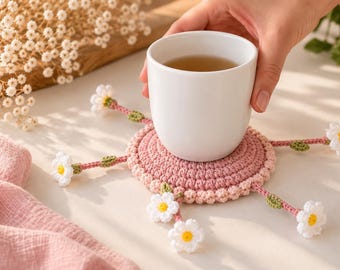 Puede incluir: Un posavasos redondo de ganchillo en color rosa empolvado con un borde texturizado, que presenta cinco tallos radiantes rematados cada uno con una pequeña flor de margarita blanca con centro amarillo y una diminuta hoja verde, bajo una taza blanca.
