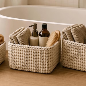 May include: Three rectangular, cream-colored woven storage baskets made of thick cord. Each basket contains folded beige fabric cloths, a wooden-handled brush, and amber or white bottles with pump dispensers, organized for bathroom storage.