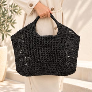 Peut inclure: Un grand sac fourre-tout noir doté d'une structure en crochet à mailles ouvertes et de deux anses intégrées. Le sac présente une base large et structurée ainsi qu'un corps souple et texturé en matériau sombre et brillant.