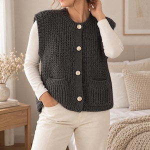 May include: A dark charcoal gray sleeveless knit vest featuring a ribbed texture, a round neckline, and four large gold-toned buttons down the front. The garment includes two patch pockets on the lower front and is worn over a long-sleeved white shirt.