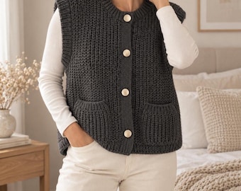 Puede incluir: Un chaleco de punto sin mangas en color gris carbón oscuro con textura acanalada, cuello redondo y cuatro botones grandes de tono dorado en la parte delantera. La prenda incluye dos bolsillos de parche y se lleva sobre una camisa blanca.