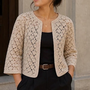 May include: A beige open-front cardigan featuring a detailed crochet lace pattern with three-quarter length sleeves, worn over a black top with black trousers and a brown leather belt with a silver buckle, creating a layered outfit.