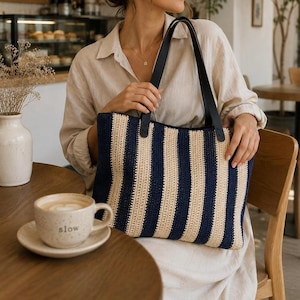May include: A rectangular tote bag featuring vertical stripes in navy blue and beige. The bag is constructed from a textured, woven material and includes two long, smooth black leather shoulder straps attached to the top edge of the bag.