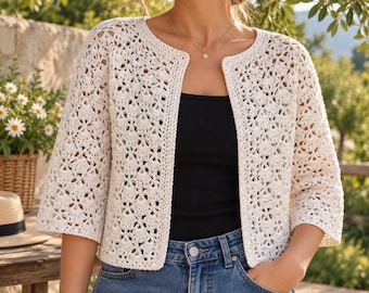 May include: An off-white, open-front crochet cardigan featuring a delicate, repeating floral-inspired pattern with three-quarter length sleeves and a round neckline, worn over a plain black top and blue denim jeans.
