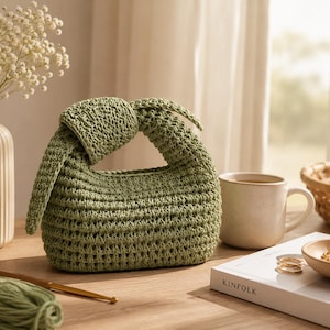 Puede incluir: Un bolso de mano de ganchillo en color verde salvia con un diseño de asa superior anudada. El bolso está construido con un patrón de punto abierto texturizado y tiene una forma redondeada y compacta. El asa se forma con un nudo superior.