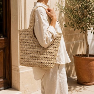 May include: A large, rectangular tote bag crafted from woven tan straw material. The bag features a consistent, open-knit texture throughout its body and includes two integrated shoulder straps that extend from the top edges of the bag.
