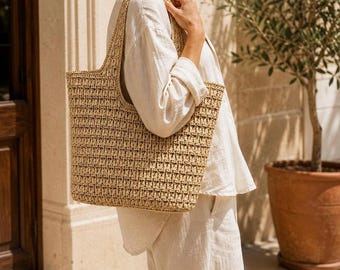 Puede incluir: Un bolso tote grande y rectangular hecho de material de paja tejida en color tostado. El bolso presenta una textura de punto abierto y consistente en todo su cuerpo e incluye dos correas integradas en los bordes superiores.