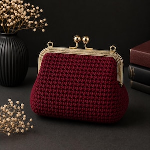 May include: A burgundy crochet clutch with a gold-toned clasp. The bag has a rounded shape and a textured surface. The clasp features two small, round accents. A black vase with dried flowers is in the background.