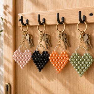 Può includere: Quattro portachiavi a forma di cuore realizzati con perle sintetiche in rosa chiaro, nero, arancione e verde salvia. Ogni portachiavi presenta un anello in metallo dorato, una catenella e chiavi argentate, appesi a ganci neri su una tavola di legno.