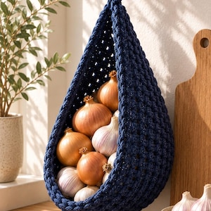 May include: A dark blue, teardrop-shaped hanging basket made of thick, braided cord. The basket is suspended from a black hook and is filled with several whole yellow onions and white garlic bulbs, showcasing a textured, open-weave construction.