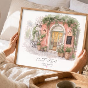 Op de afbeelding: Een ingelijste aquarelprint van een roze cafégevel met klimplanten, potplanten en terrasmeubilair. De tekst luidt "Our First Date", "Sophie & Daniel" en "September 18, 2022" met een klein rood hartje onder de datum.