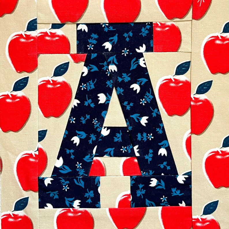 May include: A patchwork quilt block featuring a large letter A in the centre. The letter is made from dark blue fabric with a white floral pattern. The background of the block is a beige fabric with red apple shapes.