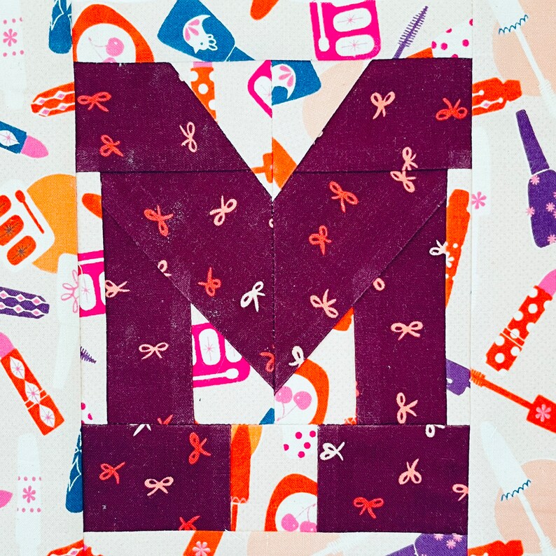 May include: A purple fabric block with a large 'M' shape created by sewing together smaller fabric squares. The fabric squares have a white background with a repeating pattern of pink bows.