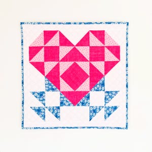 May include: A pink and blue patchwork quilt with a heart design. The heart is made of pink and white fabric with a diamond pattern in the center. The quilt is surrounded by a blue and white border.